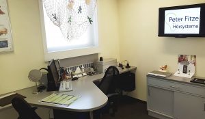 Modernes Hörakustiker-Büro mit Schreibtisch, Computer und Bildschirm „Peter Fitze Hörsysteme“.